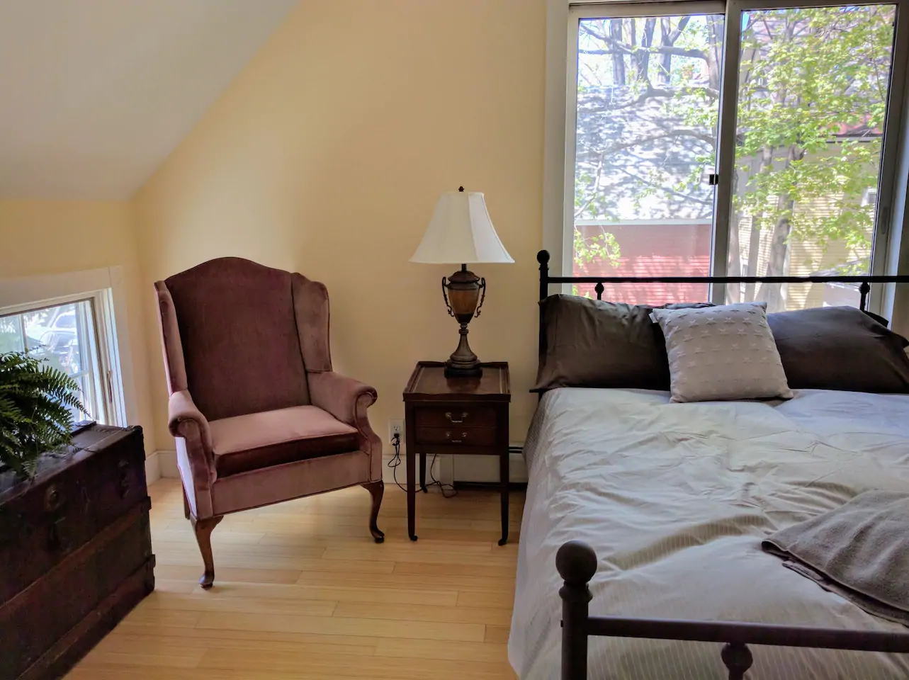 A bedroom with a bed made with cream bedding. A window is above the bed looking out to trees. A red chair sits next to the bed and sidetable with a light.