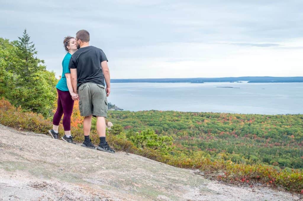 A man and a woman kissing in front of a grassy area with a body of water in the distance