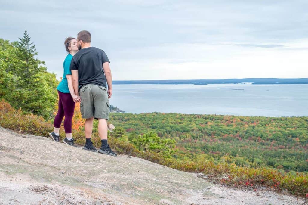 Couple kissing while standing on a rock. In the distance, trees lead up to the water under a grey sky during a great time to visit Acadia National Park