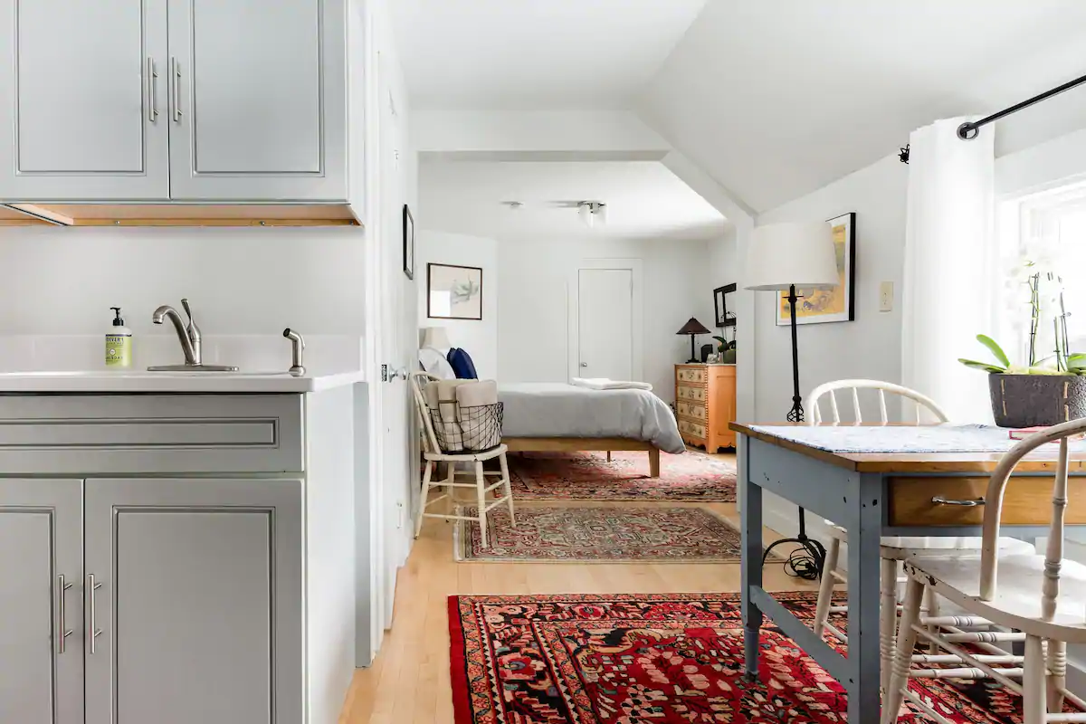 View from a kitchen of a bed with grey bedding in a studio apartment with white walls. To the right, near the kitchen, is a grey table with white chairs.