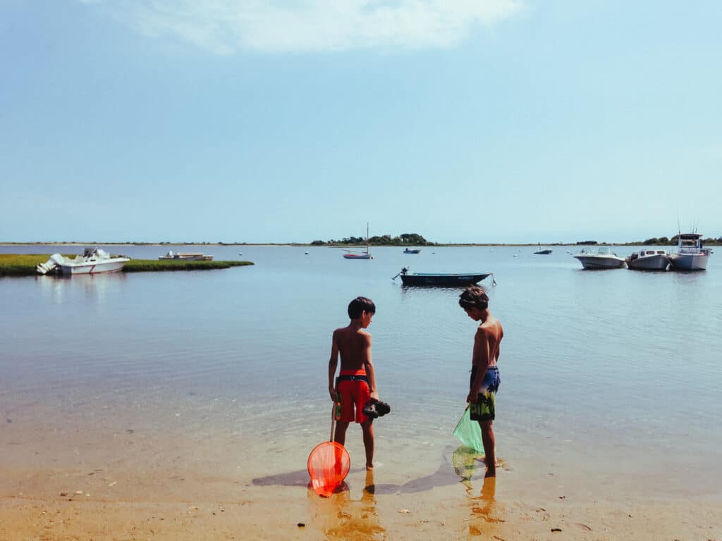 Two children standing on the edge of the ocean holding nets in a popular New England family vacation spot. Boats float on the water in the distance.