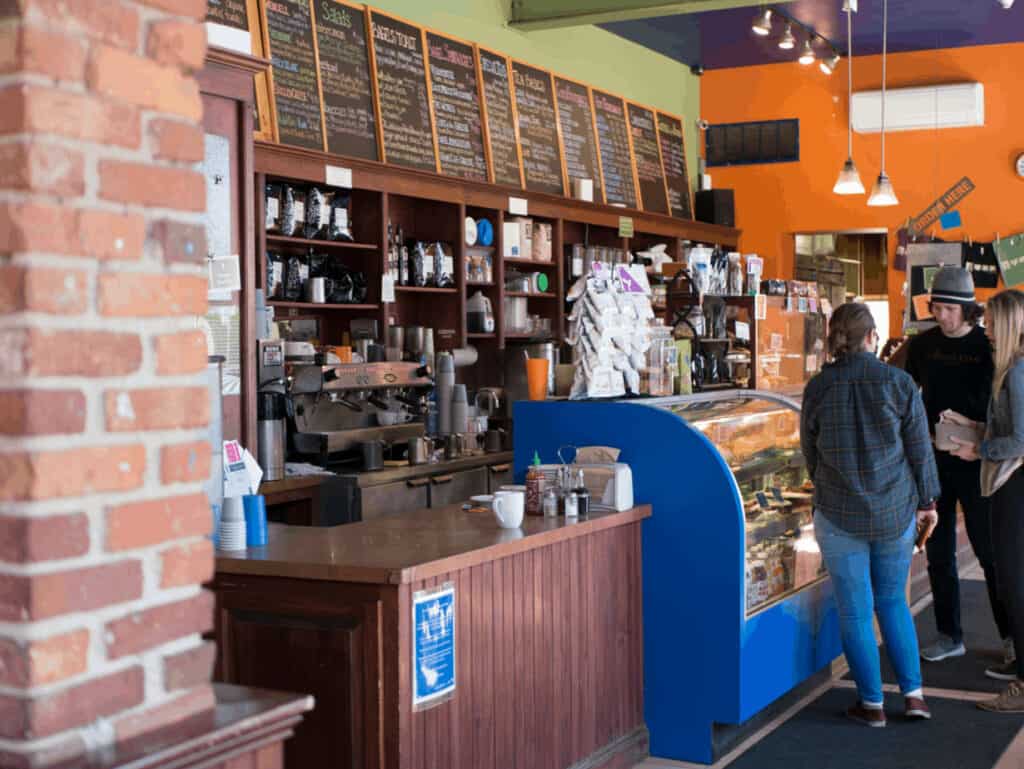 Cafe barista area, with coffee lining the walls and a blue glass case showing pastries. A woman stands in front of the glass case at a Northampton, Massachusetts coffee shop.