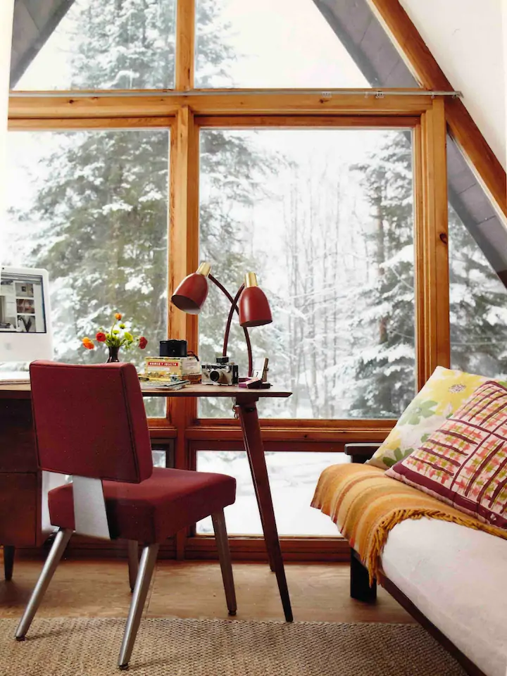 A red chair facing a table and a large wall of windows looking outside toward the woods