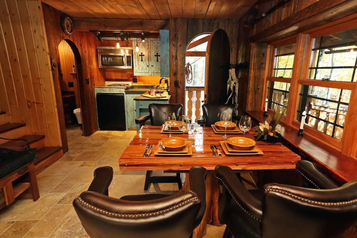 Interior of a kitchen and dining area with dark wood paneling on the walls and ceiling. Leather chairs sit around a wooden table with place settings in the forefront. The background has a door ajar, leading to the outdoors, and a blue kitchen area.