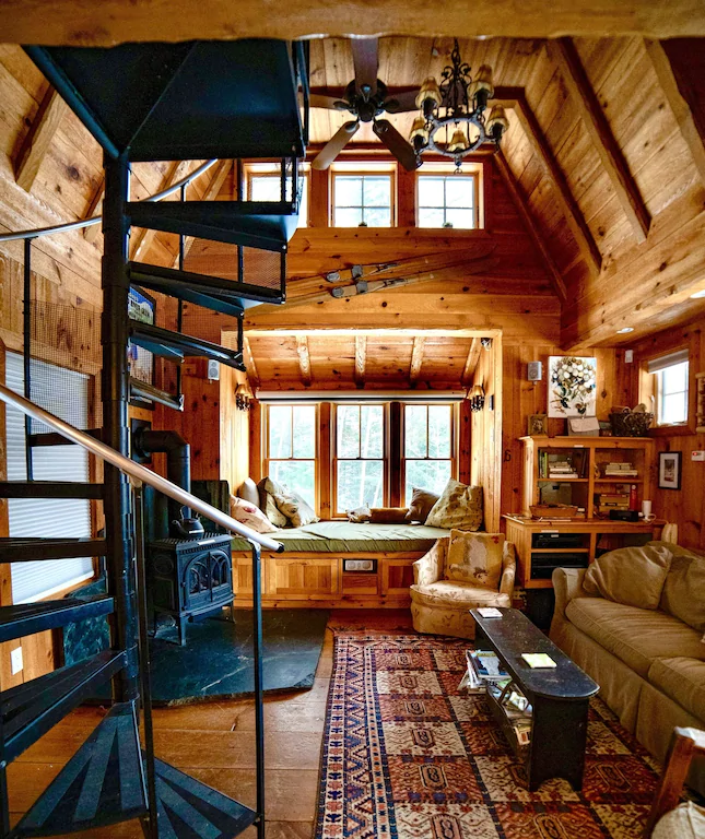 A spiral staircase on the left in a wood paneled room with windows in a Vermont cabin
