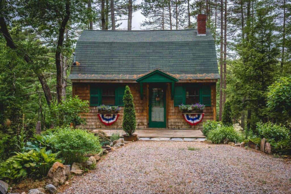 A wooden cabin with green shutters sits alone surrounded by tall trees.