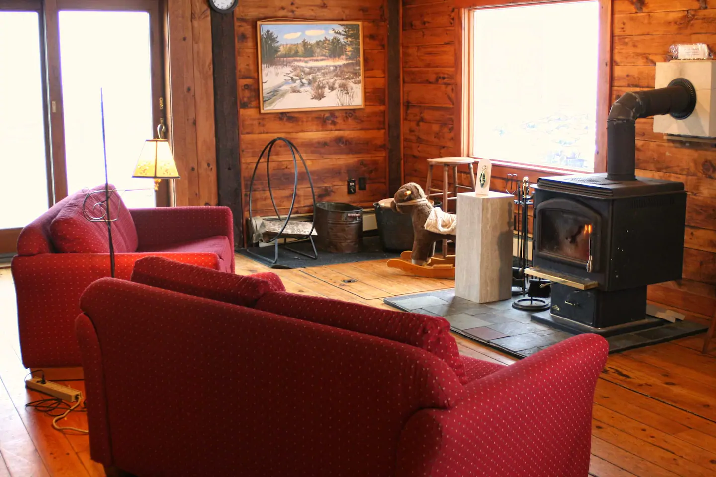 Interior of a cabin living room. Two red couches sit around a pellet stove. Light seeps through a window behind the stove.
