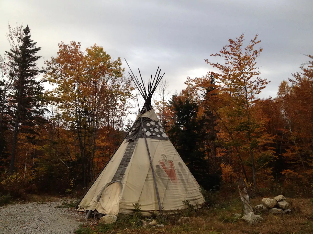 A brown and cream tipi sits surrounded by fall foliage as the sky turns dark grey.