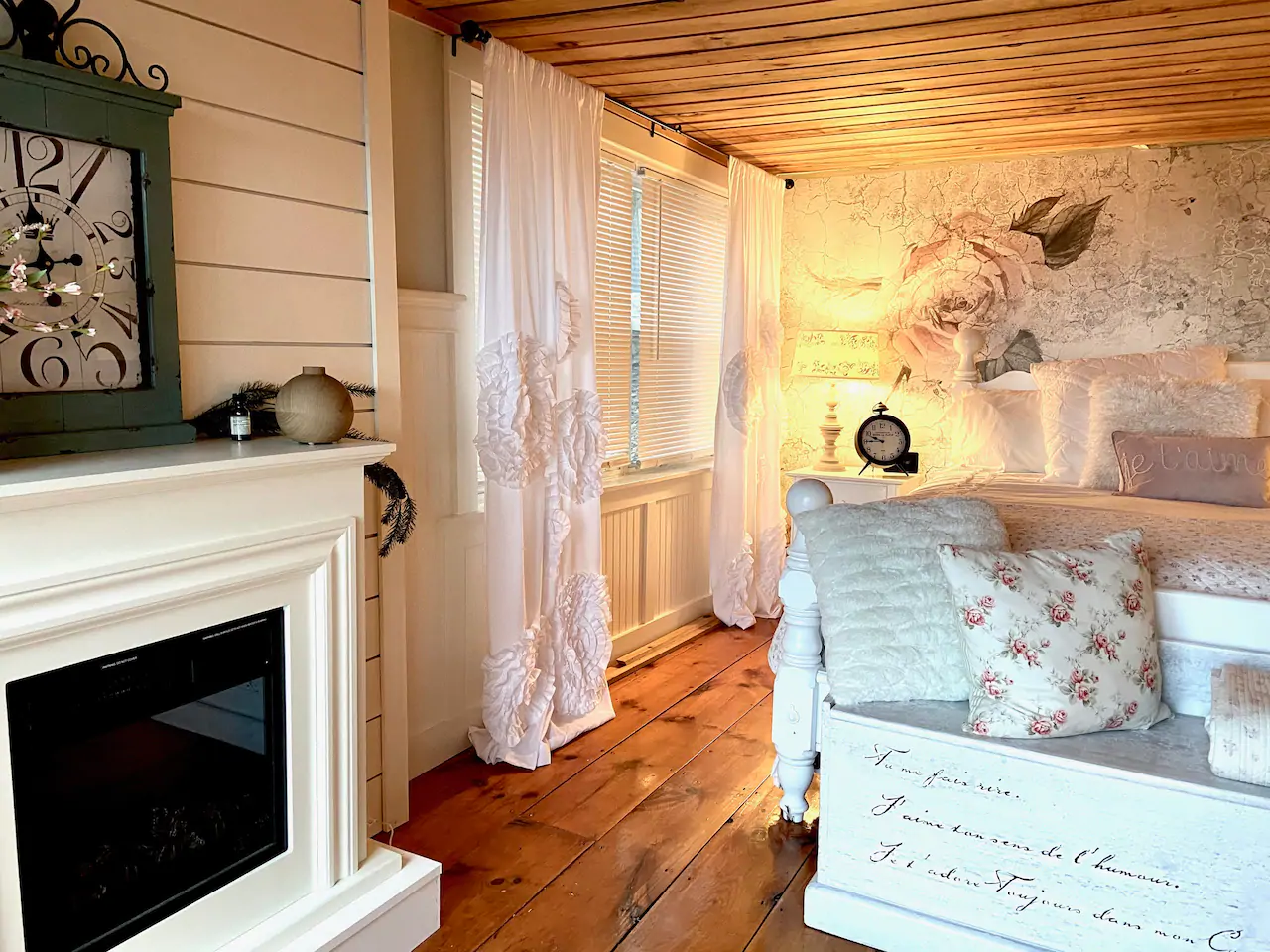 Interior of a bedroom with a cottage core theme. To the left is a white fireplace and a window with its shades closed. To the right is a plush bed with white bedding.
