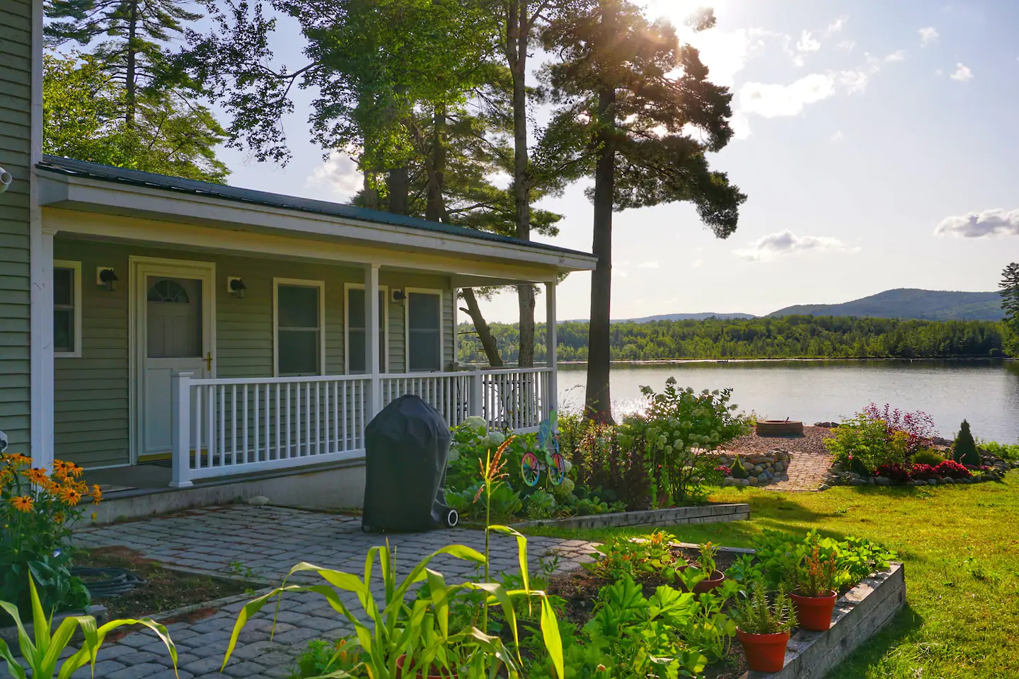 A green home sits surrounded by tall trees, a garden in the front, and the lake in the distance at a vacation rental in New Hampshire.