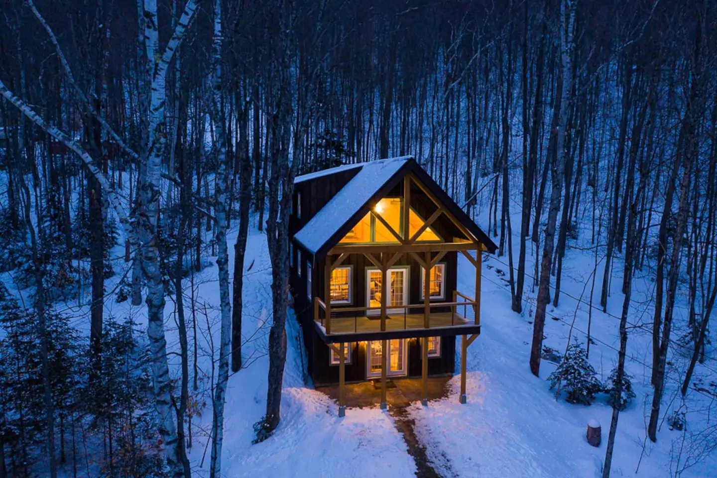 A two story chalet with interior lights on in the middle of the snowy woods at night