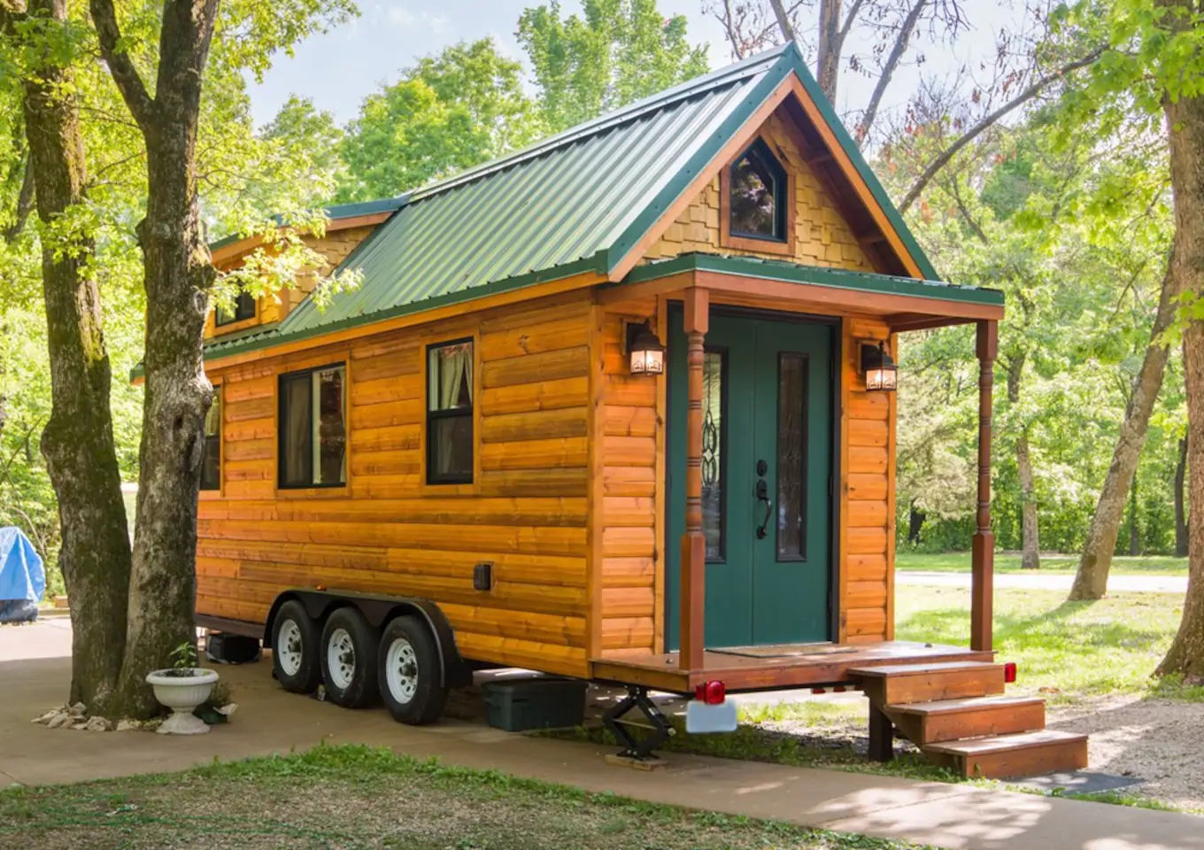 A small tiny house made of wood with a green double door. Stairs lead up to the home, which is on wheels and is surrounded by a plush forest.