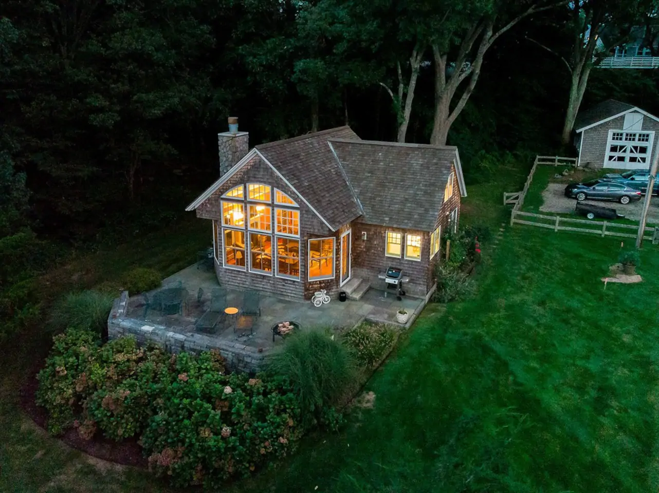 Classic New England style home is lit up in the evening with big windows that showcase a living room. The home is surrounded by greenery and trees.