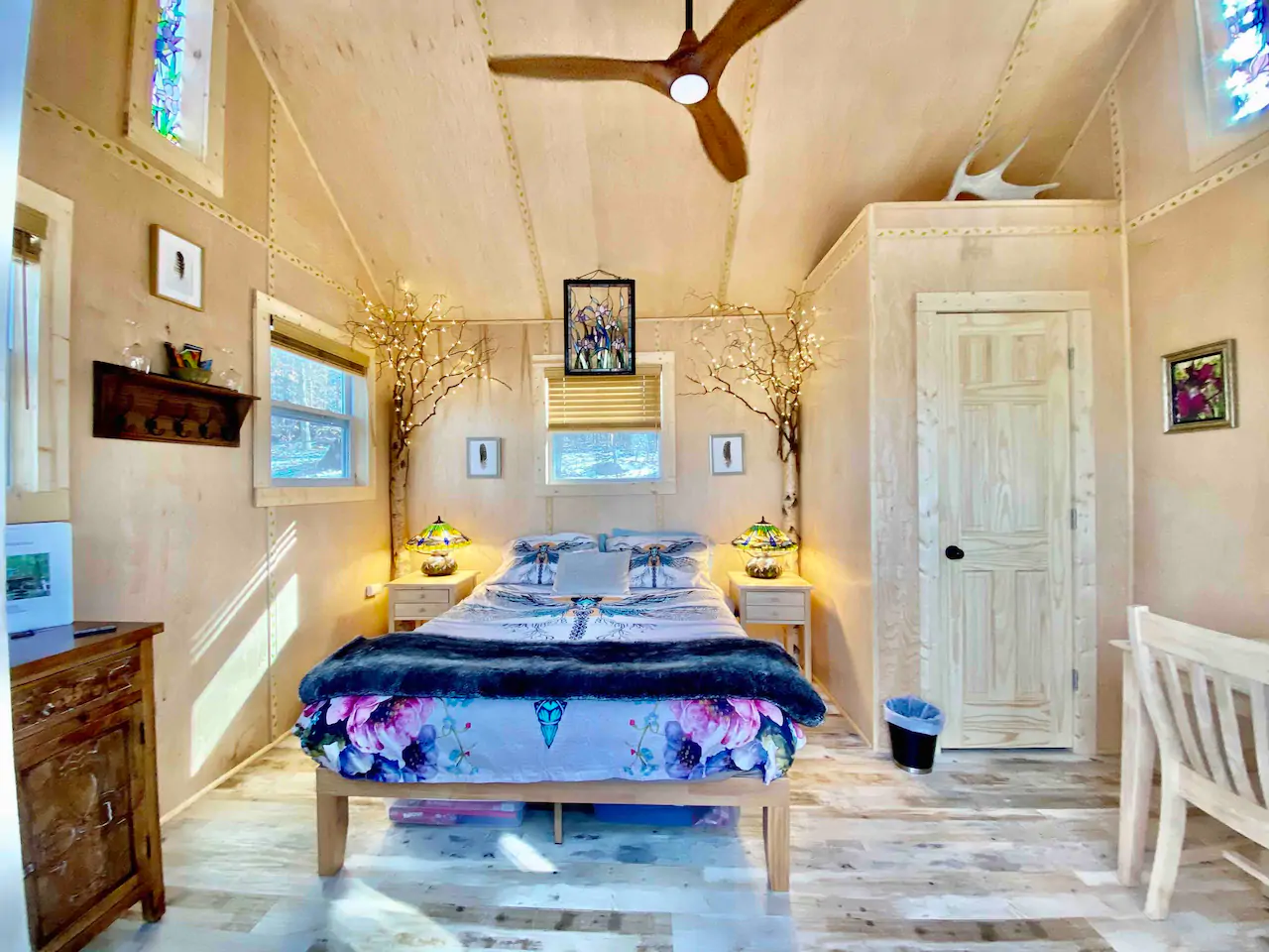 A bed is made with purple and blue bedding. Above the bed is a wooden ceiling fan.