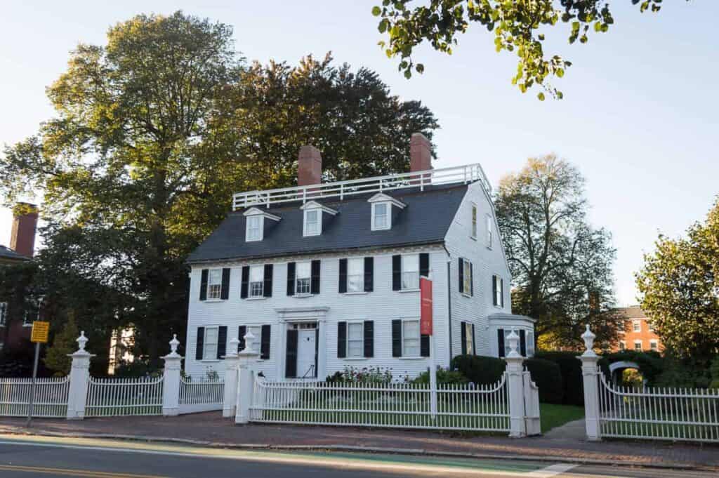 A large white historic home looms large surrounded by trees and a white wrought iron fence under a clear sky in Massachusetts.