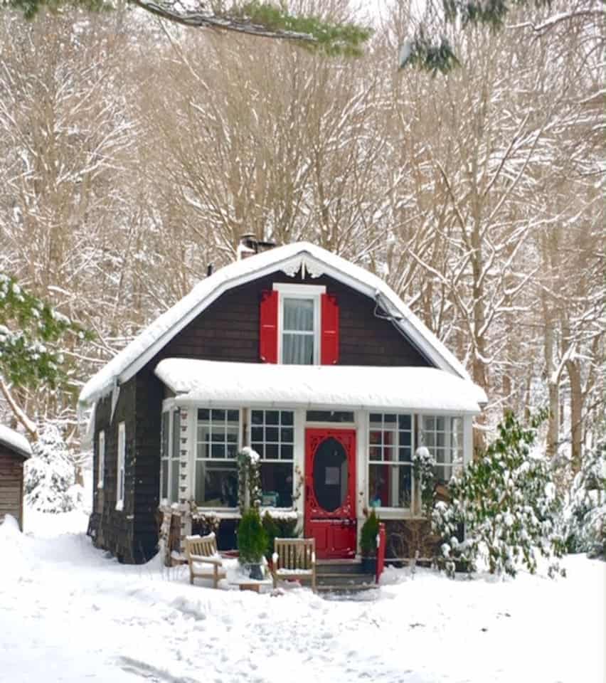 A black and white house with a red door and red shutters in the middle of a snowy forest.