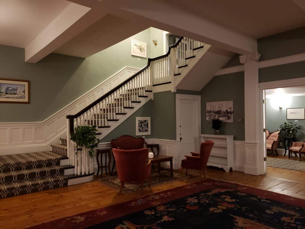 Interior of an inn with green walls. A stairwell leads upstairs. A few red chairs are located in the corner by a table.