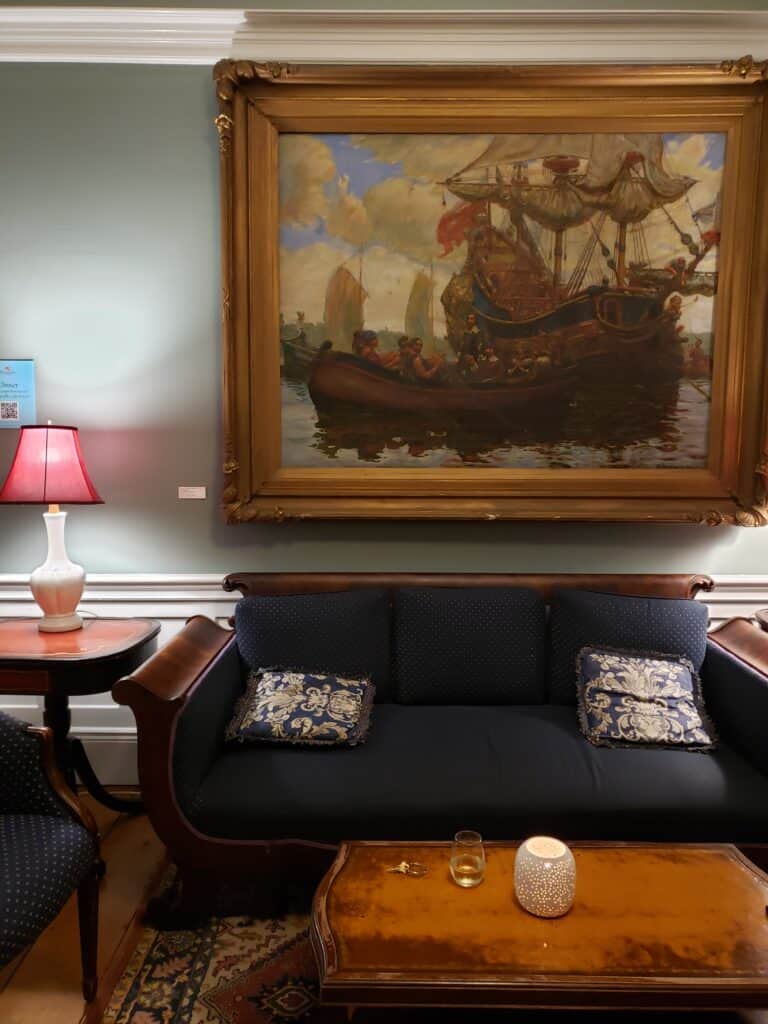 A living room filled with wooden furniture, most notably a couch with blue upholstery. Above the couch is a painting of a ship.