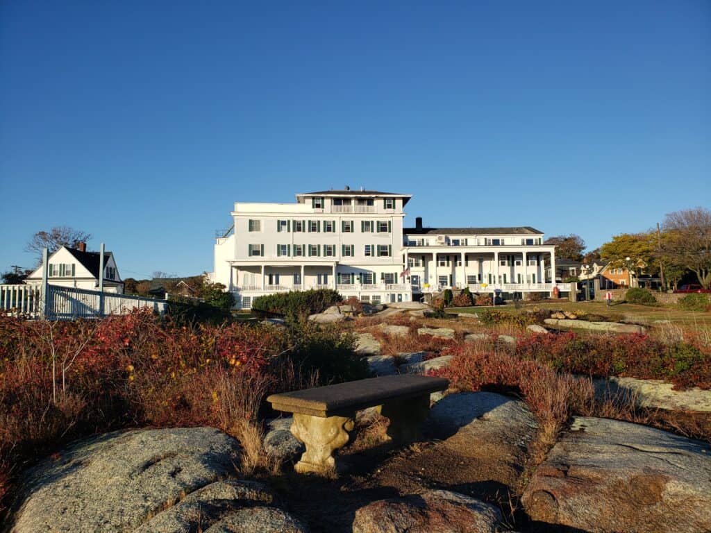 A historic white inn sits under a blue sunny sky. In the foreground is a stone bench on a rocky edge.