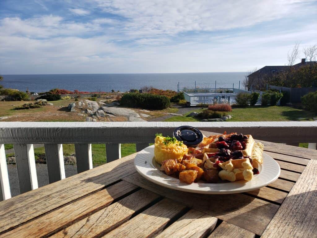 Breakfast with waffles and potatoes sitting on a white plate on a wooden table. The distance features a view of the water.