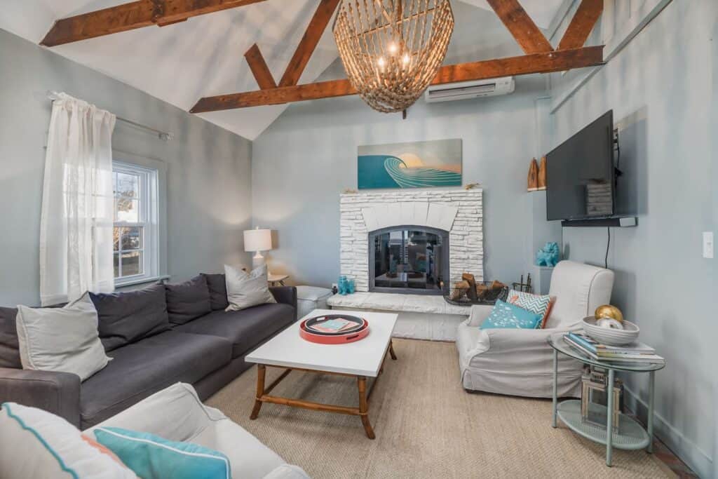 Living room with high ceiling with wooden beams and chandelier. A dark gray couch is on the left, with a coffee table in front. At the far side of the room is a white fireplace.