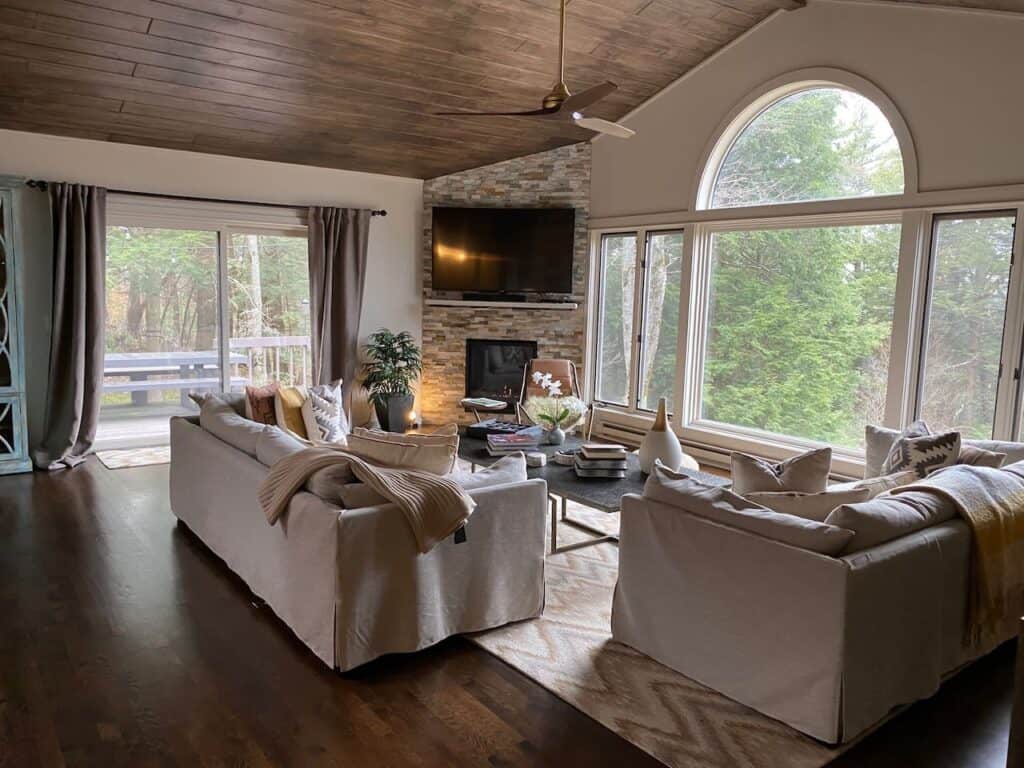 A white sofa and chair facing a tv on the stone wall, a sliding glass door and a large picture window looking out at the green trees.