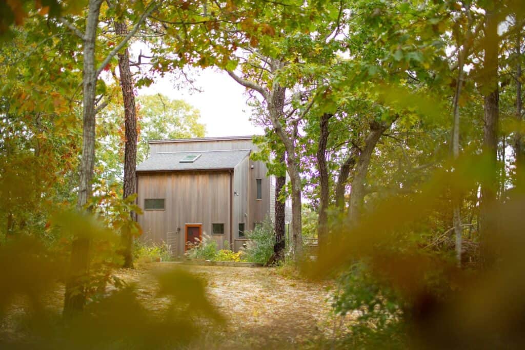 Grey wooden house with a large brown door and surrounded by trees.