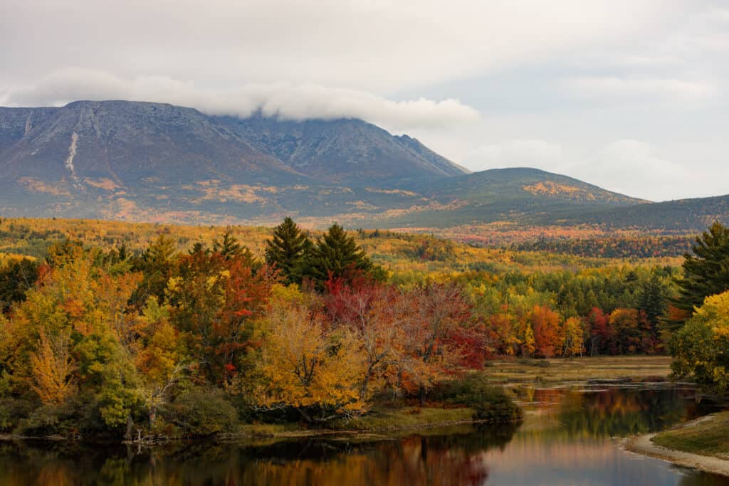 Dense fall foliage by a river extends out to a nearby mountain under cloudy skies on a Maine road trip