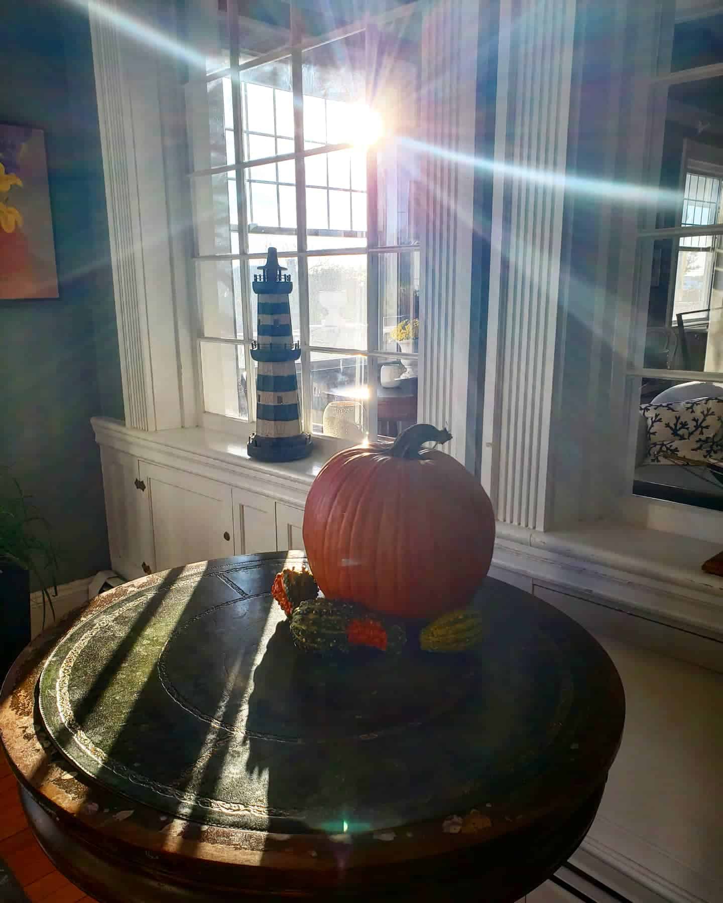An orange pumpkin with other fall gourds sits on a circular table. Behind the pumpkin, the sun peeks through the window.