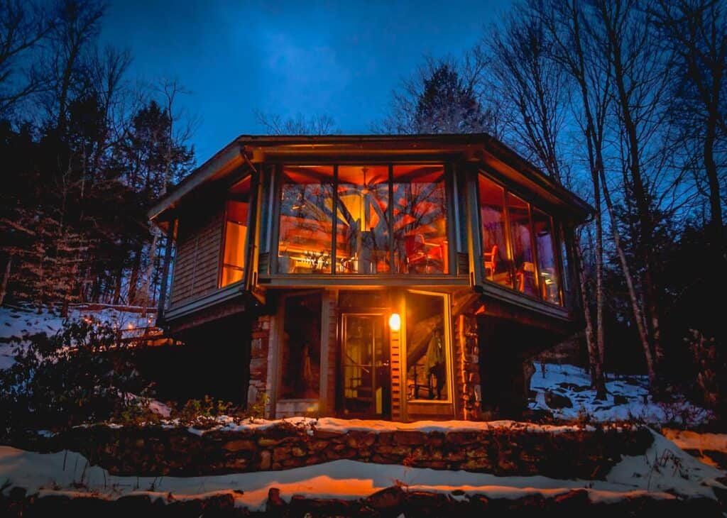 A beautiful octagonal 2 story New England Airbnb with windows all around showing the lighted interior inside at dusk 