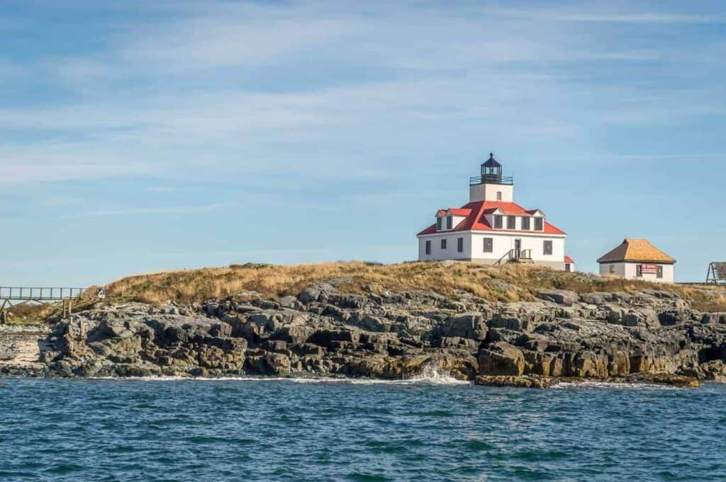 A red and white Maine lighthouse sits atop a rocky cliff with calm seas in front and light blue skies overhead.