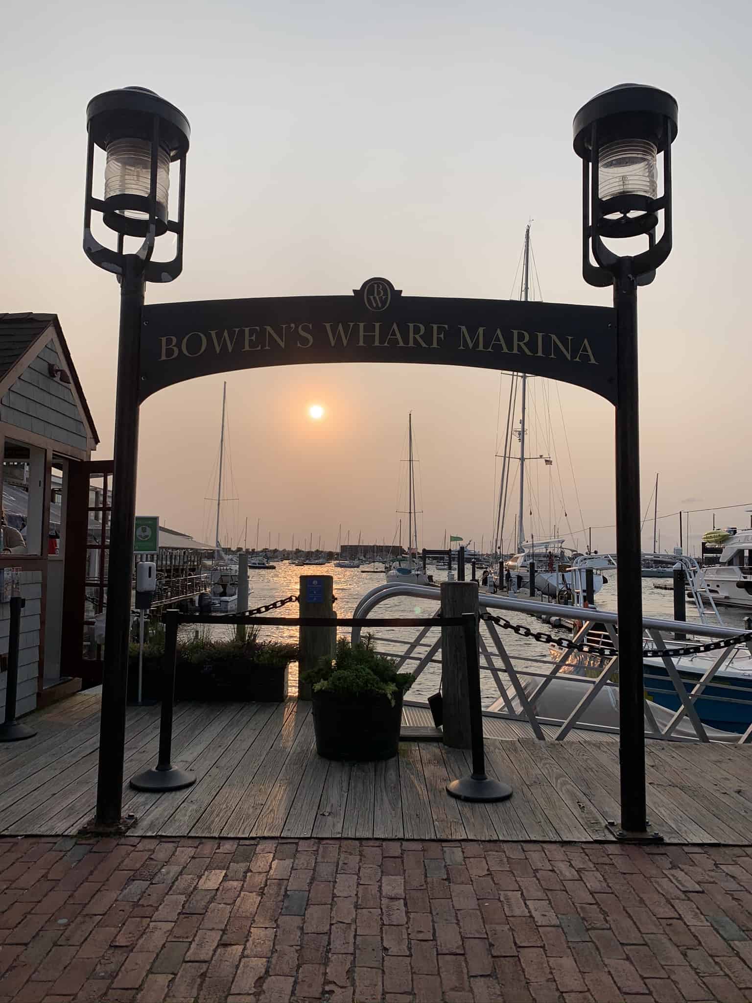 An archway at a dock with boats and a walkway with a sunrise in the distance