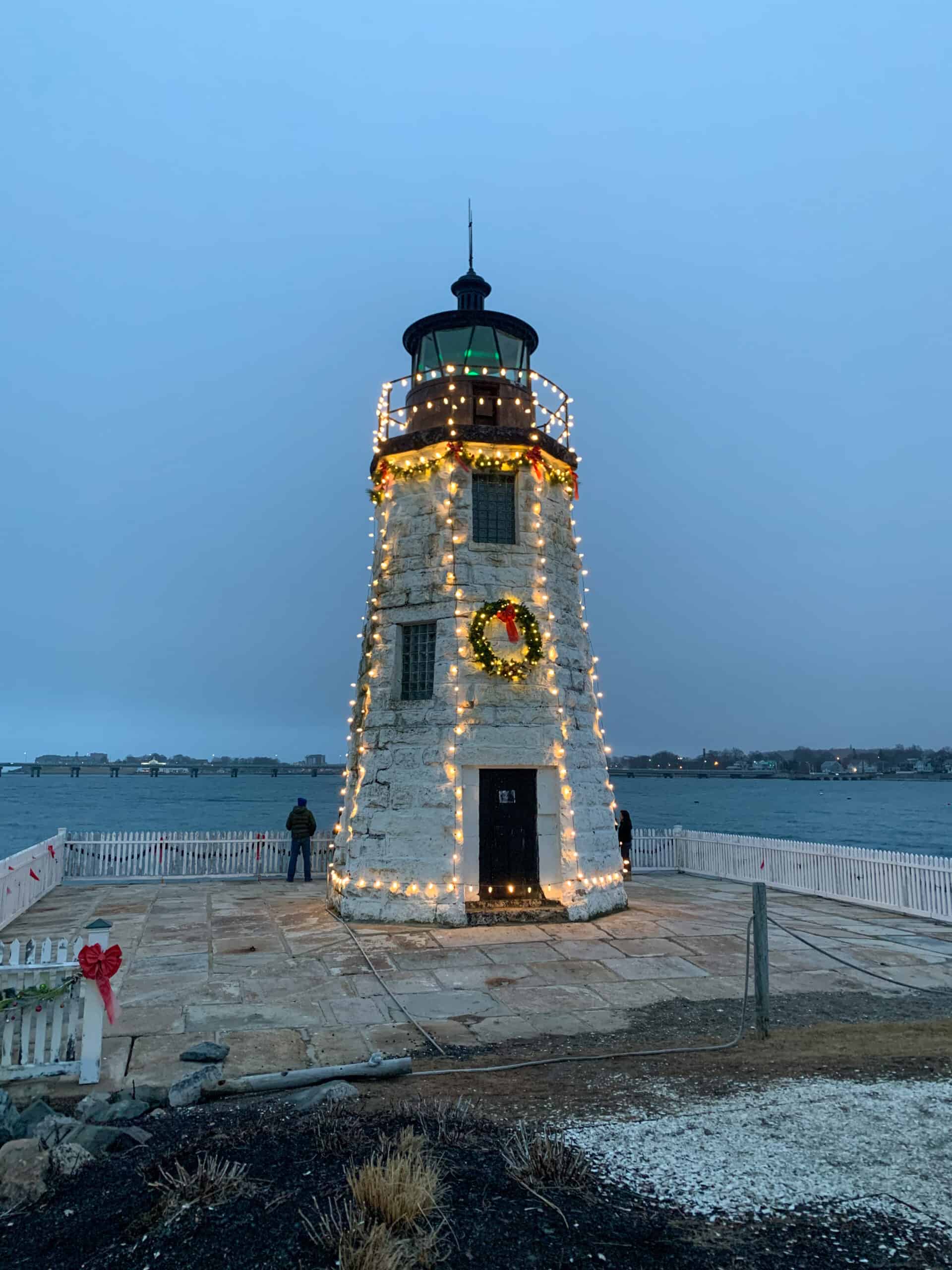 A white lighthouse with a black top decorated with white holiday lights and a wreath