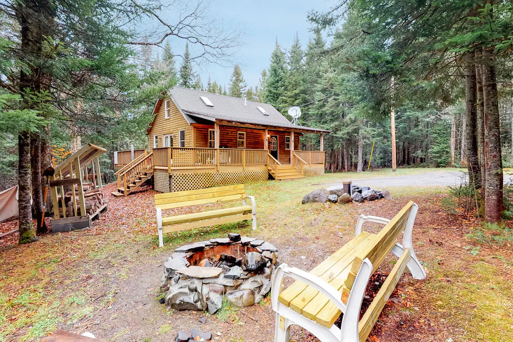 Two wooden benches facing a fire pit with a log cabin in the distance