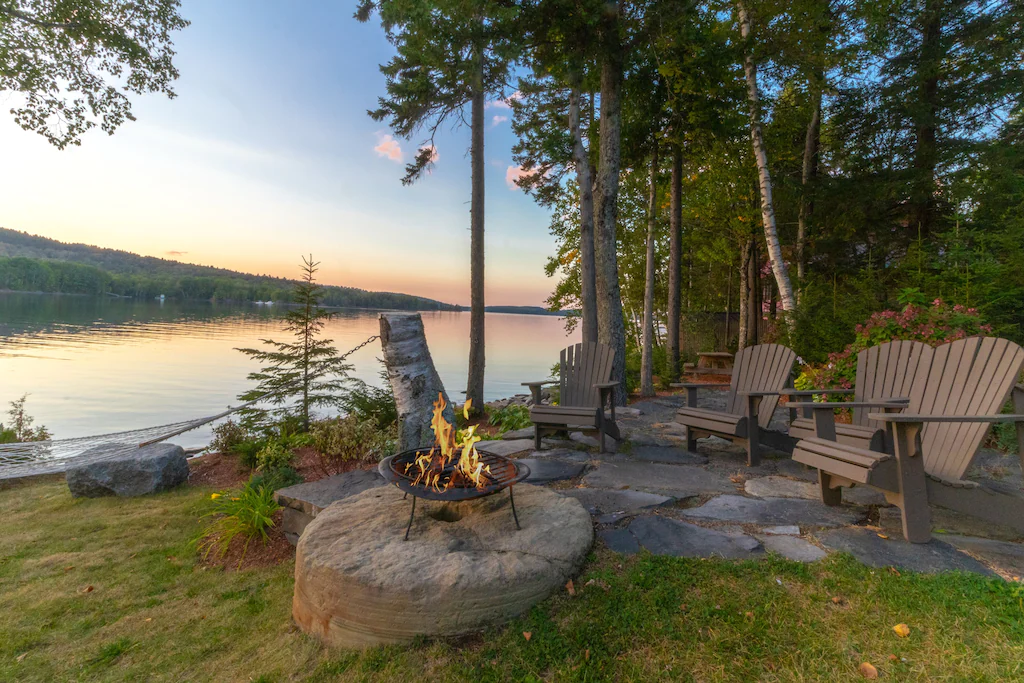 Adirondack chairs surrounding a fire pit next to a body of water at sunset