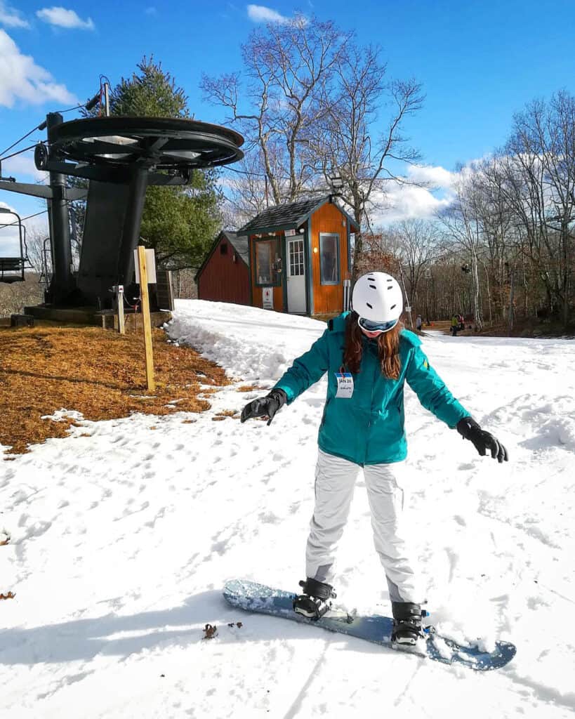 Woman wearing a teal winter coat and white snow pants standing on a snowboard in the snow. She\'s also wearing black gloves and a white helmet.