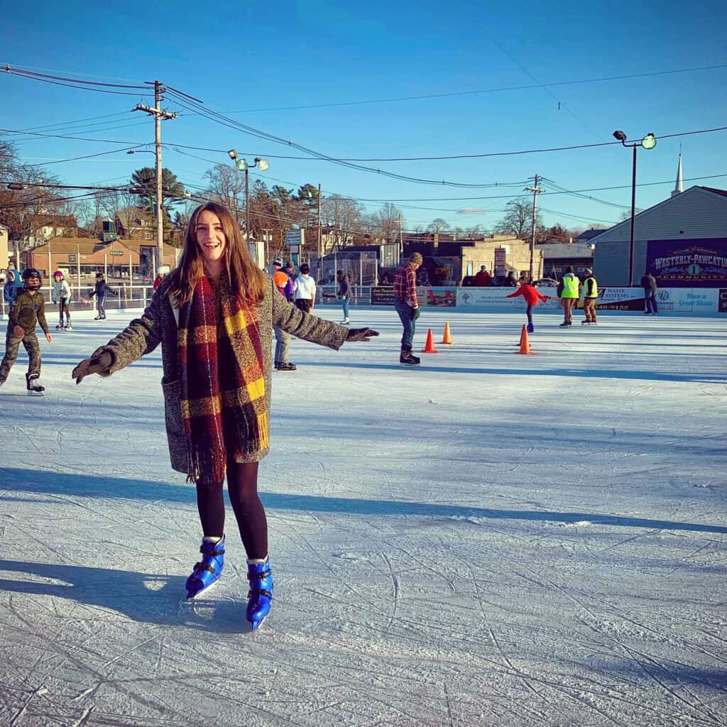 Woman in winter clothing and a red and yellow scarf smiling wide while ice skating outdoors.