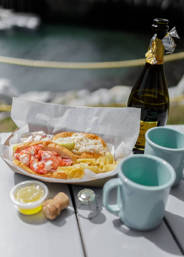 Lobster roll siting on a plate next to two light blue mugs and a bottle of wine.