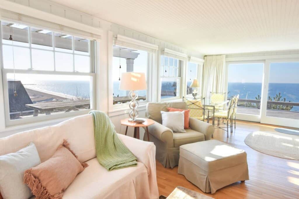 Interior of a living room with lots of natural lighting. The foreground features a light pink couch with a green blanket laying on it. The ocean can be seen out the window.