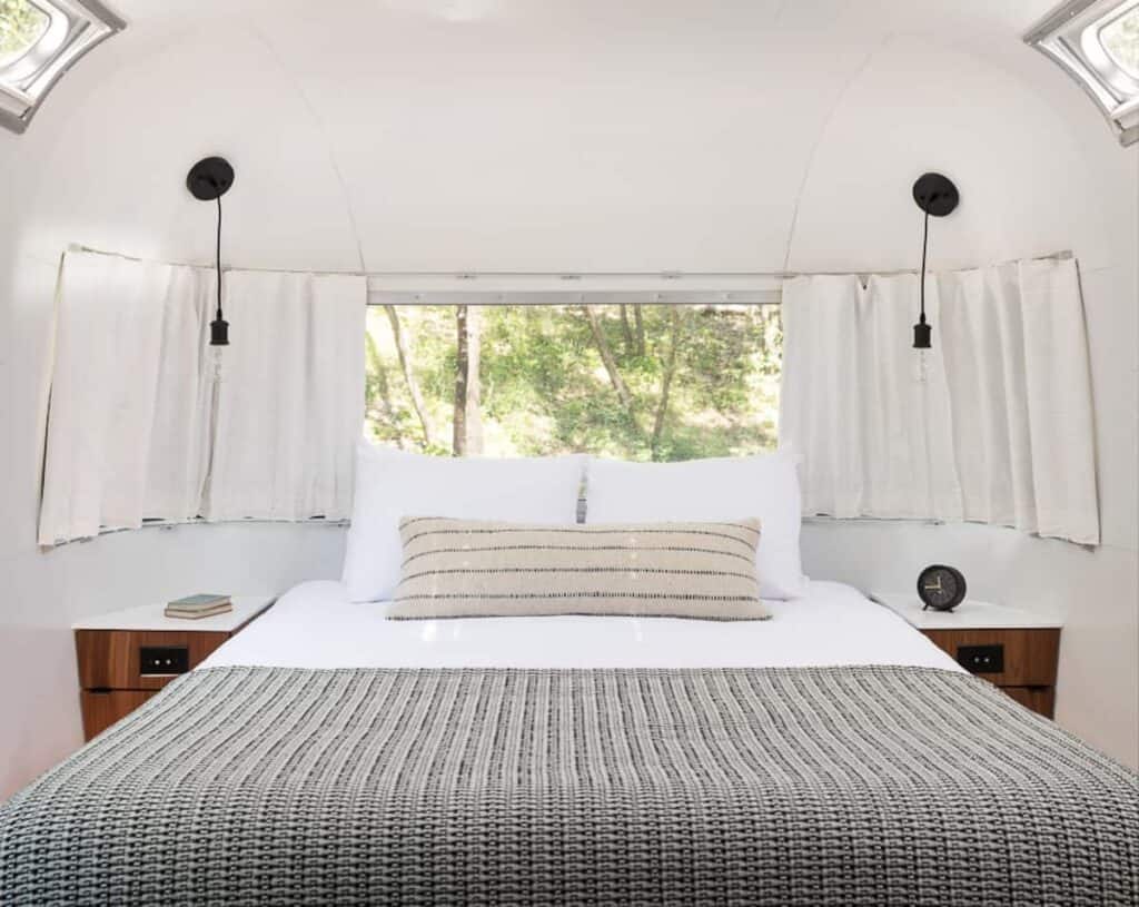 Interior of a hotel room with a well-made bed with grey bedding. A window behind the bed shows a green forest.