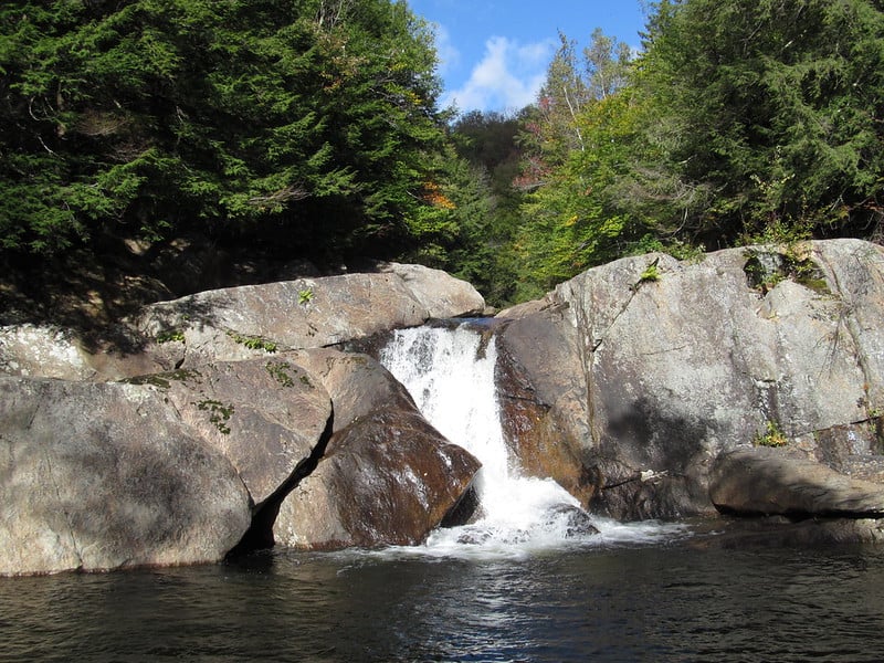 Big waterfall splitting a large rock in half as it falls into the water under a blue sky by a forest.