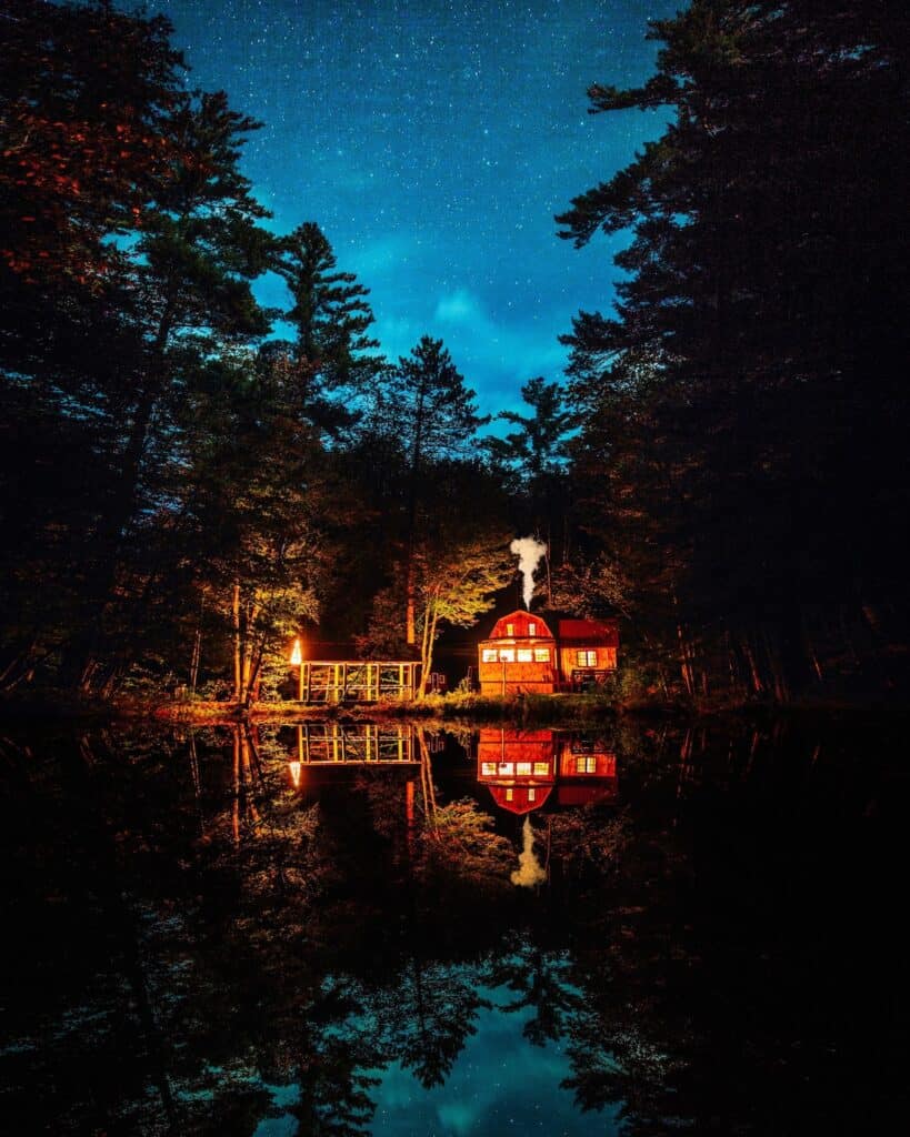 A large cabin in the woods at dusk with the lights reflecting in the water