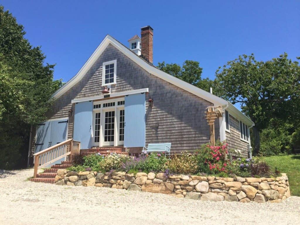 New England cottage with blue accents. A stone brick wall lines the edge of the home near the coast.