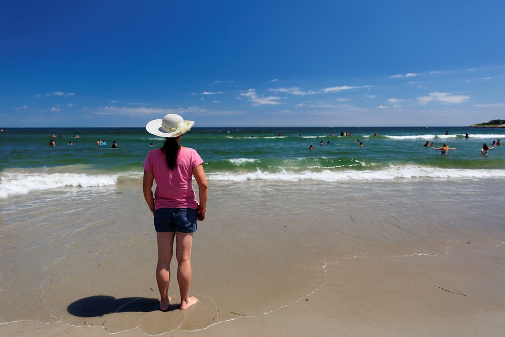 A person with a pink shirt, jean shorts, and white hat standing with their feet in the water looking out to the ocean under the blue sky at one of the best beaches in Rhode Island.