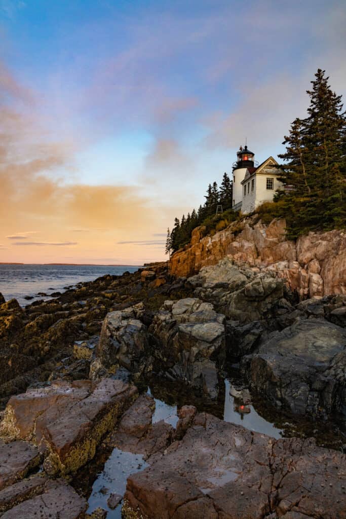 A lighthouse sits perched on a rocky cliff by the water, as the sun rises and creates an orange and blue sky.