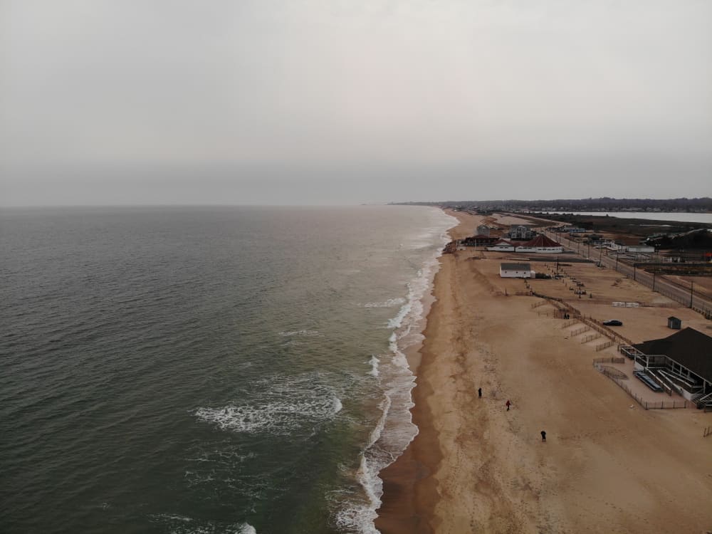 Aerial view of the coast as the waves crash against the sand. In the distance, white buildings can be found near the coast. People are walking in the sand in the foreground at one of the most popular Rhode Island beaches.