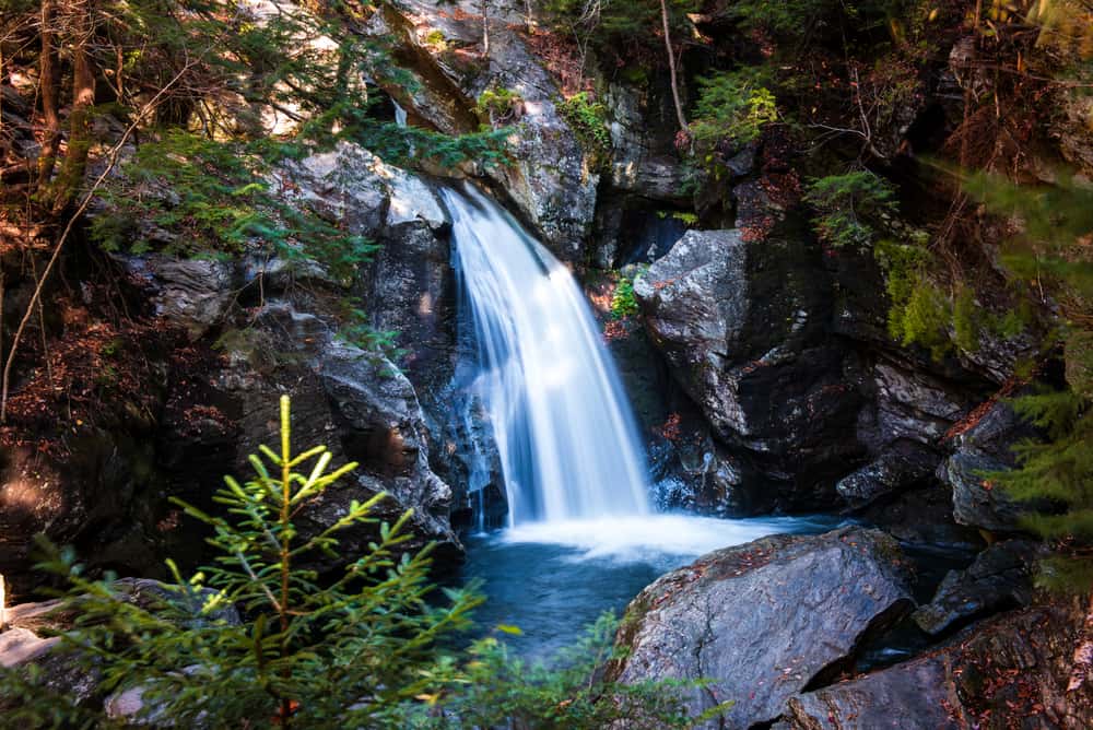 Blue waterfall with mist falling into a pond by wet rocks.