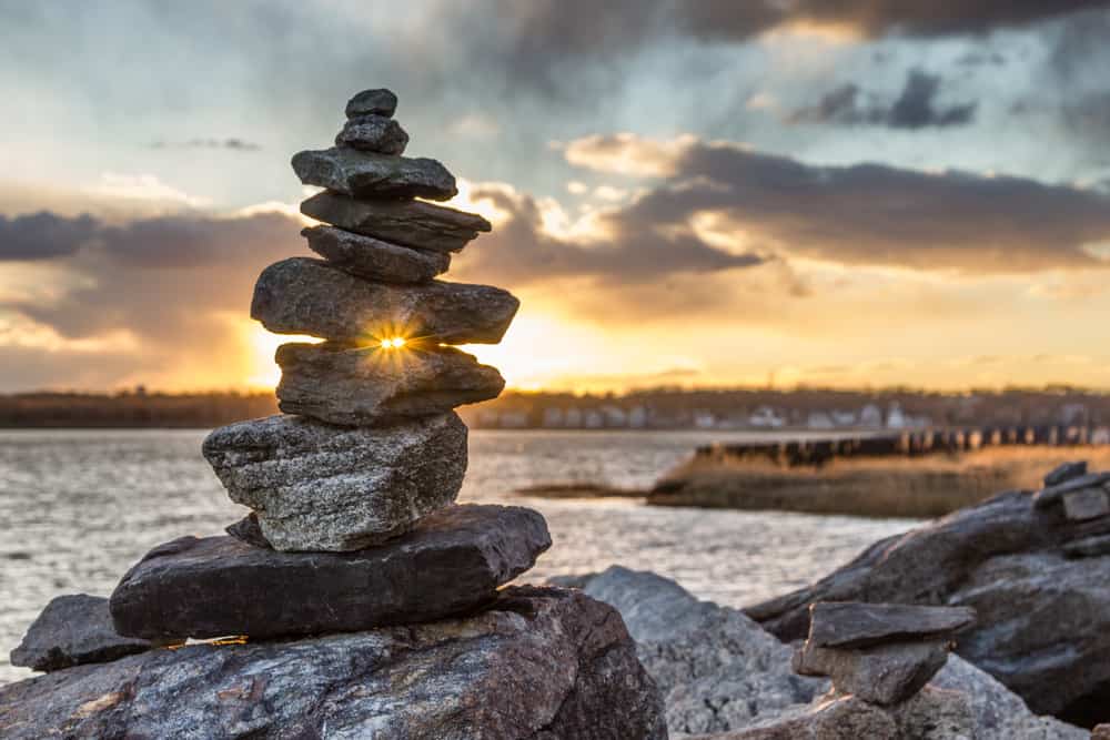 Close up of a stack of rocks with the sun peeking through two of the rocks. The ocean can be seen behind the rocks, under a sunset sky.
