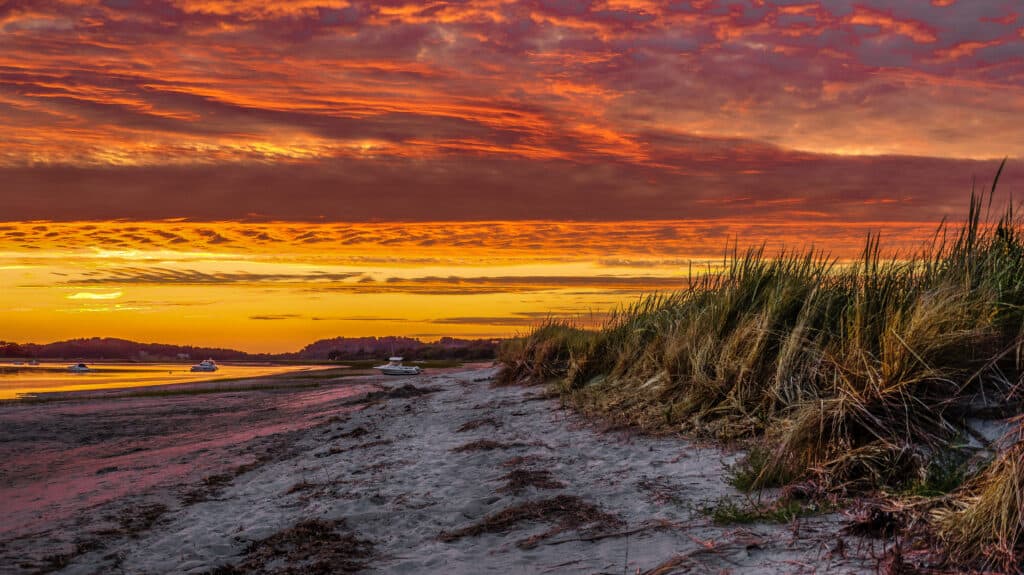 One of the best beaches North Shore MA has to offer is pictured as the sun sets with a fiery red sky overhead and a calm beach with sea grass on one side and a still sea on the other