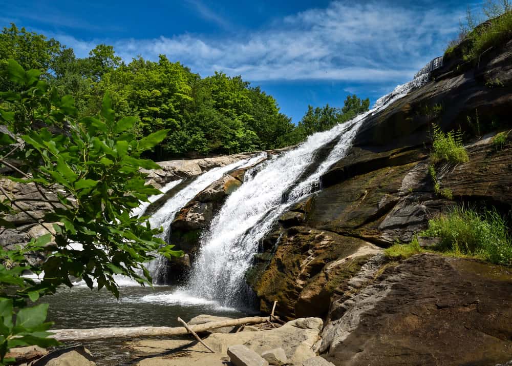 Three Connecticut waterfalls cascade down a cliffside into a body of water under a blue sunny sky.